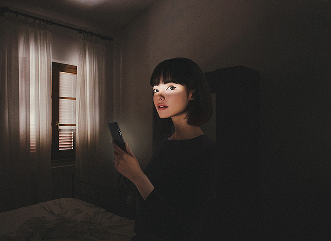 Woman lit by phone screen in dark bedroom.