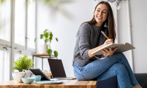 Image of a woman sitting at an office desk, looking outside while writing in a notebook, representing a thoughtful approach to understanding freelancers' needs.