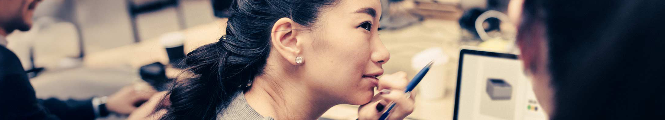 An office setting with a young woman attentively listening while holding a pen near her face. She is seated at a workstation with a laptop displaying a data visualization.