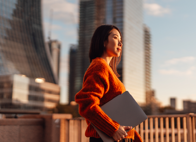 Smiling woman wearing glasses and a blue shirt working on a laptop by a window.