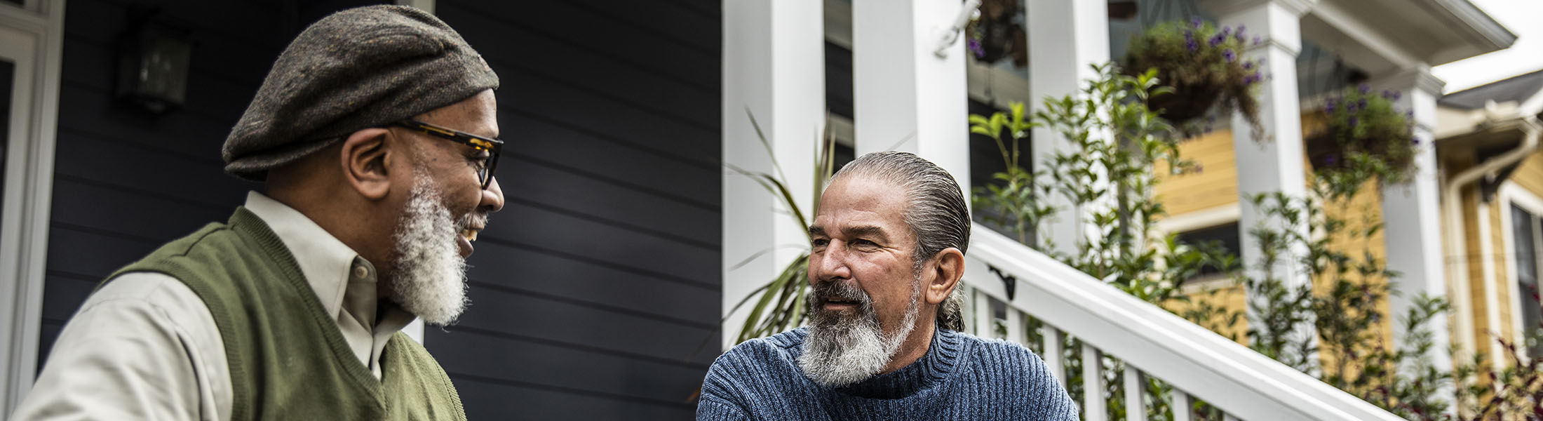 Two older men sitting on a porch, smiling and talking in front of suburban homes.