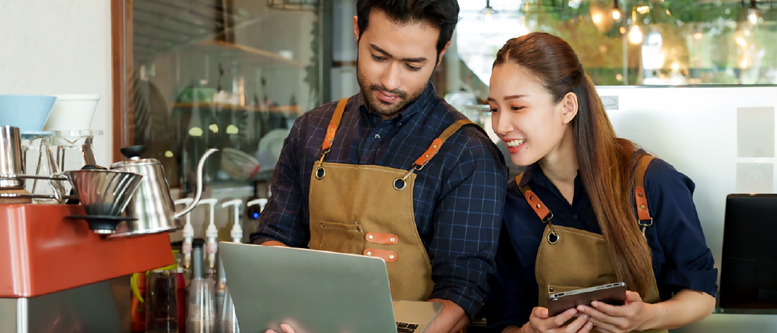 Two coffee shop workers in aprons smiling while looking at a laptop behind a counter.